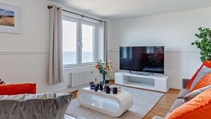Moderni olohuone, jossa on suuri litteänäyttöinen televisio, valkoinen sohvapöytä ja oransseilla koristetyynyillä varustettu sohva.