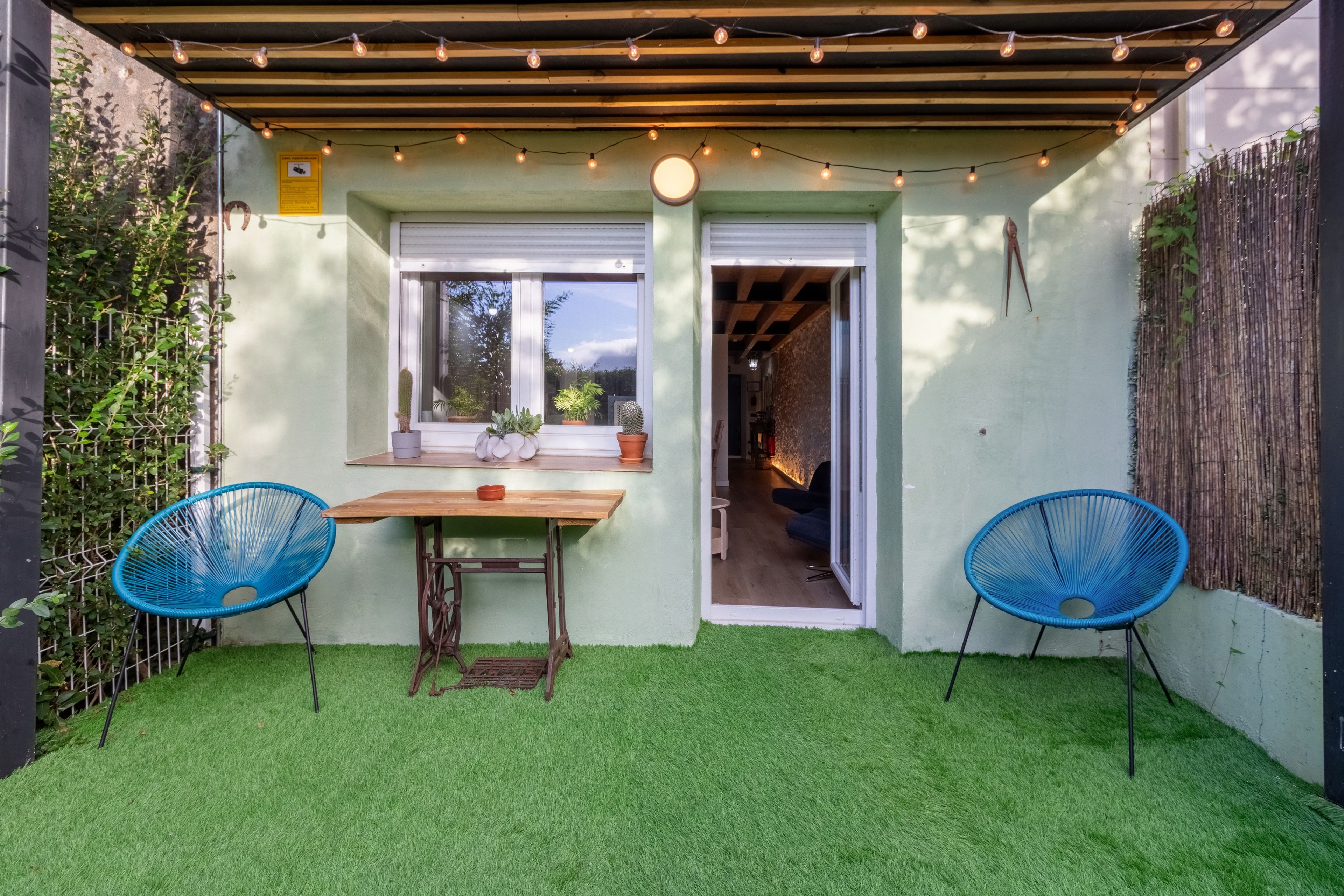 Un patio con césped artificial, una mesa de madera, dos sillas azules y una guirnalda de luces.
