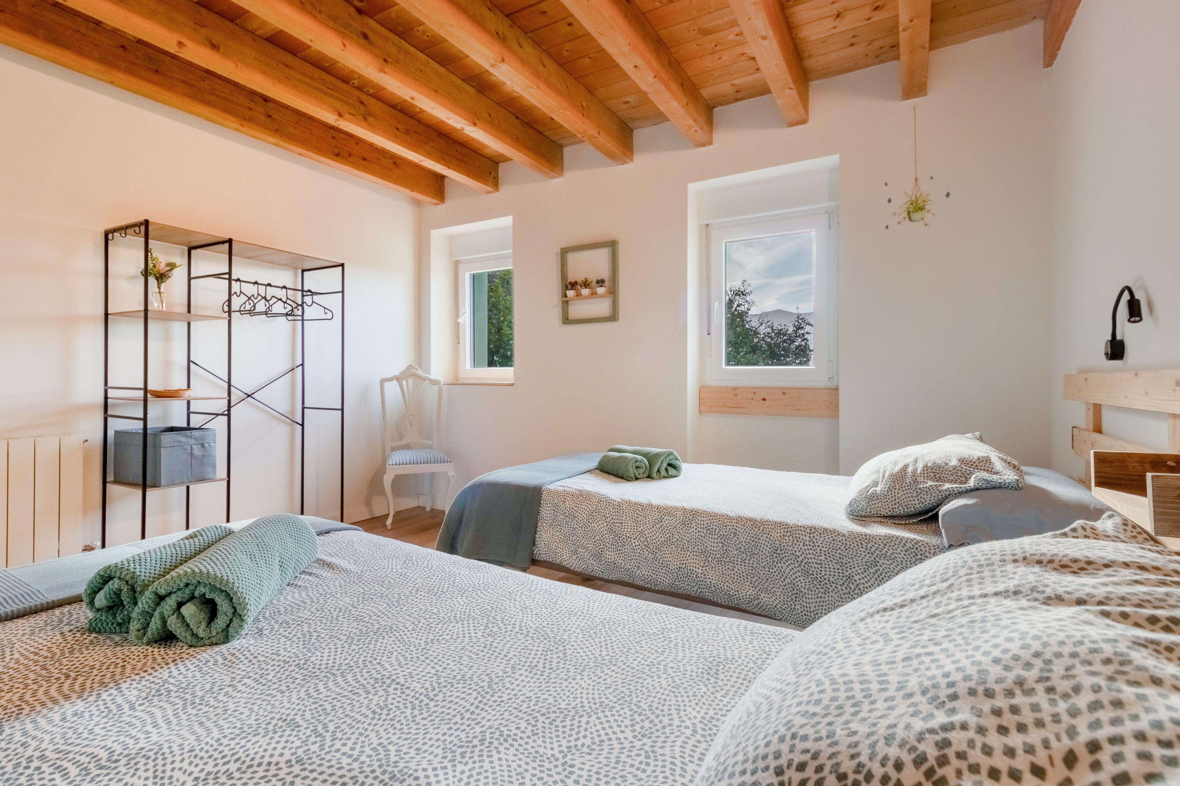 Una habitación con dos camas, techo de madera, ventana con vistas y una estantería metálica con ropa.