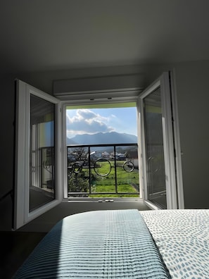 Habitació amb una finestra gran, un llit i vistes a una muntanya i verdura.