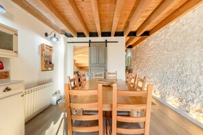 A dining area with a wooden table and chairs, a white wall with a textured section, and a ceiling with exposed wooden beams.