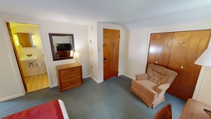 A hotel room with a bed, a wooden dresser, a chair, a wardrobe, a mirror, and a bathroom with a sink and toilet.