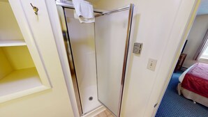 A room with a shower, a bed with a red cover, a white towel hanging, and a small shelf.
