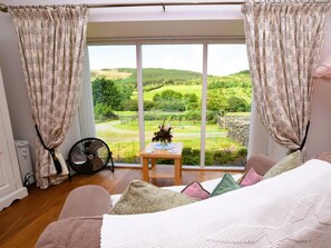 Ein Schlafzimmer mit großem Fenster, einem Ventilator, einem kleinen Tisch mit einer Vase und einem Bett mit Kissen.