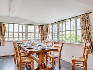 A dining room with a wooden table set for six, surrounded by wicker chairs, large windows with curtains, and a view of the outside. - 3 Bed in St. Columb (oc-82405) (Ruthvoes)