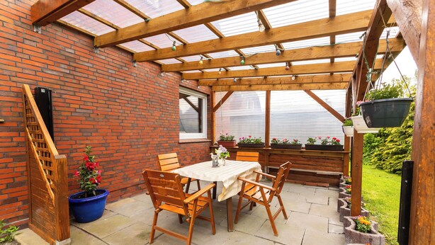 Eine überdachte Terrasse mit Ziegelwand, Holztisch und -stühlen sowie Topfpflanzen.