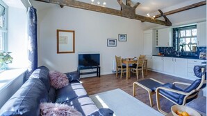 Una sala de estar con sofá, televisor, mesa de comedor y sillas. Cuenta con una zona de cocina con armarios y ventana.