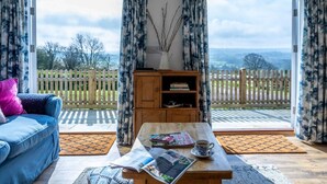 Ein Zimmer mit einem blauen Sofa, einem Holztisch mit einer Zeitschrift und einer Tasse, und Blick auf einen weißen Zaun und Grünflächen draußen.