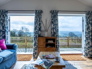 A room with a blue sofa, a wooden table with a magazine and a cup, and a view of a white picket fence and greenery outside. - 1 Bed in Axminster (oc-82604) (Tytherleigh)