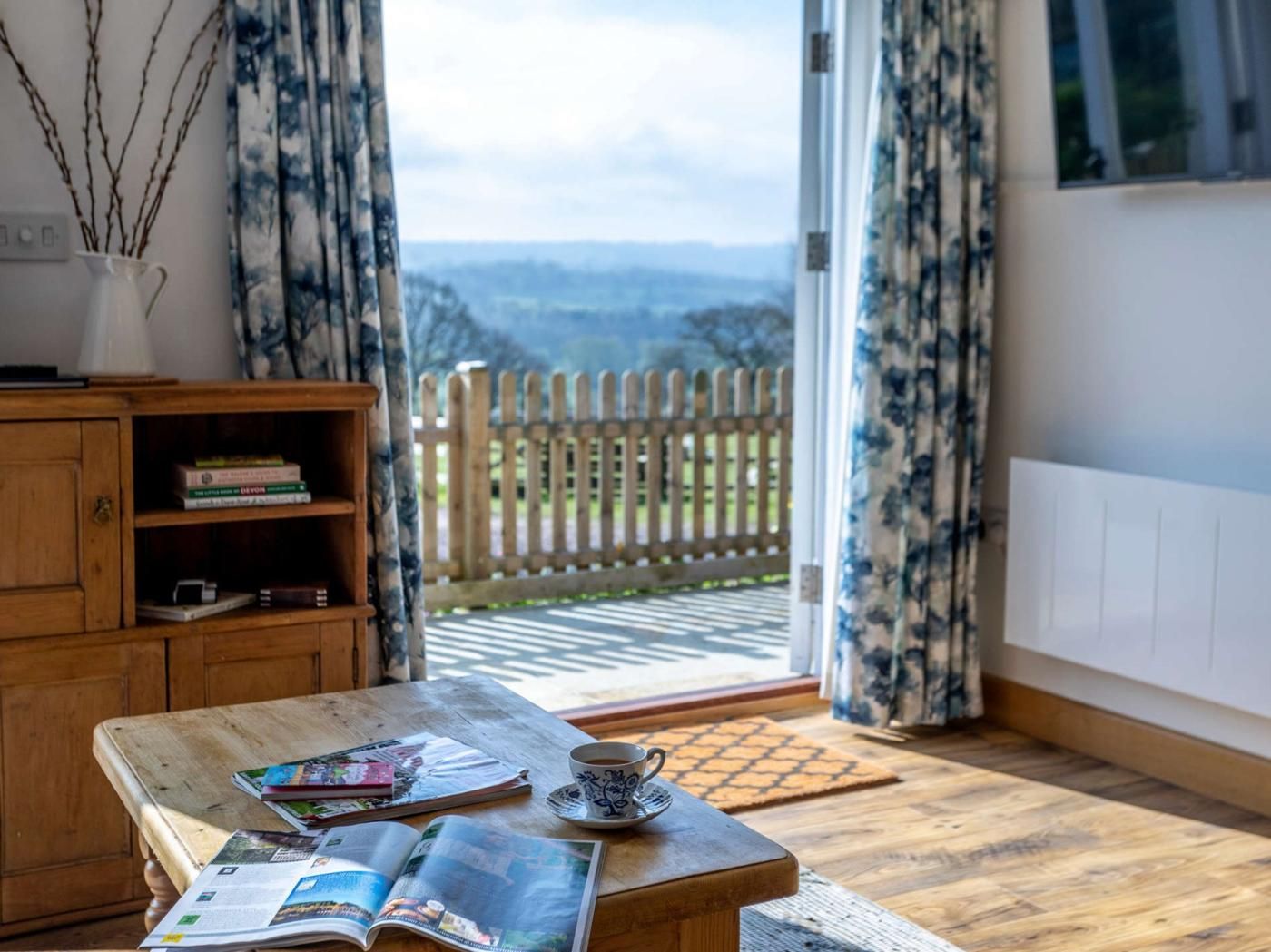 A room with a wooden table, books, a cup, and a view of a wooden fence and landscape through a glass door.