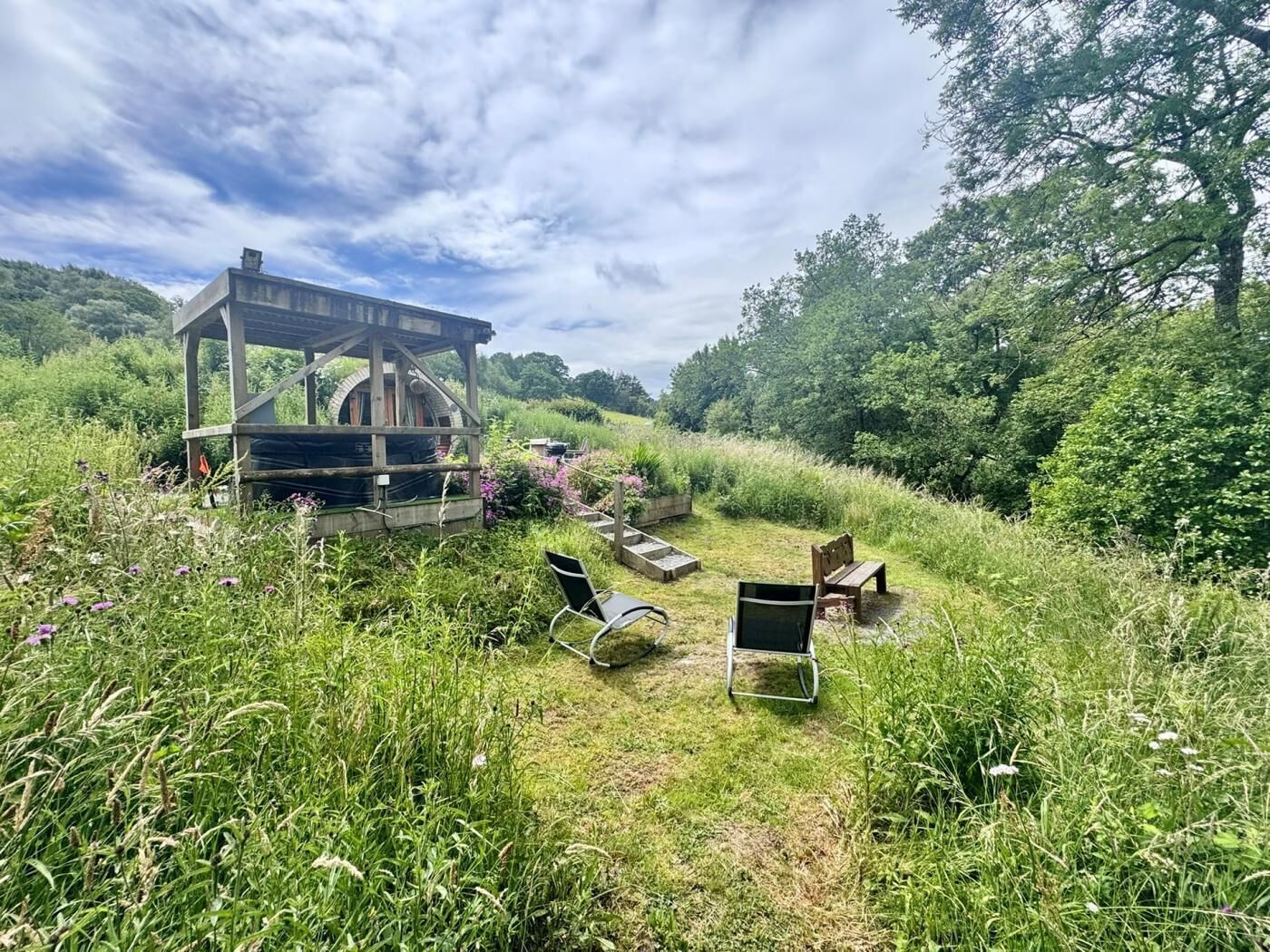 Een rustieke houten constructie met een rond raam, omgeven door een grasveld met enkele stoelen en een bankje.