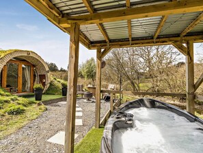 A wooden outdoor seating area with a hot tub, a covered patio, and a rustic building with a circular window.