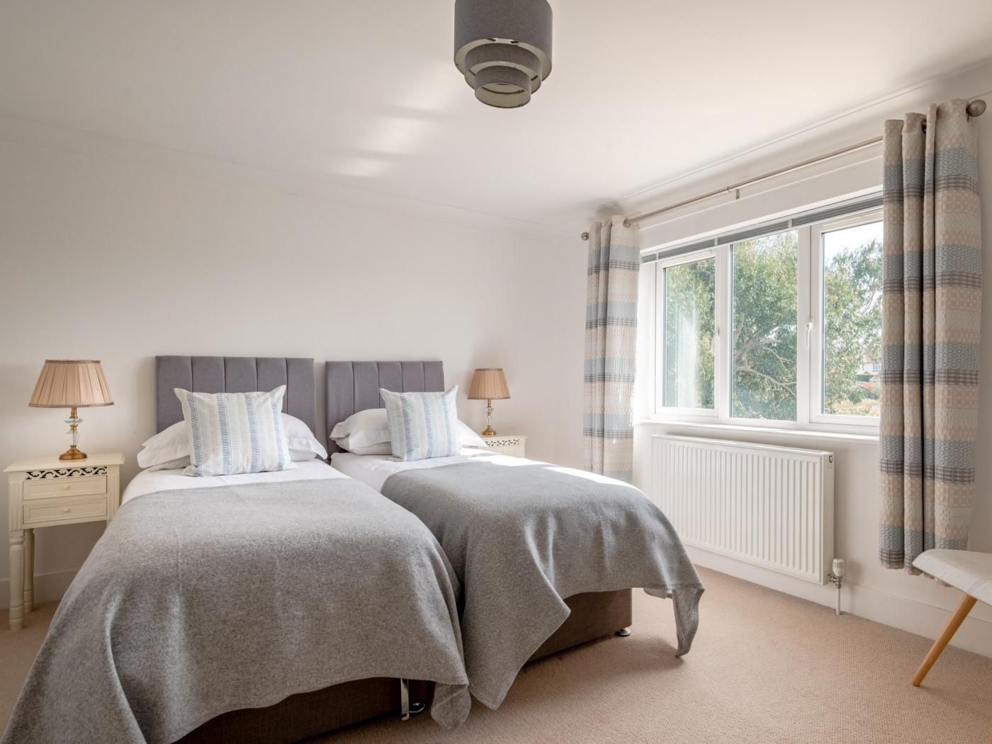 A bedroom with two beds, a window with curtains, a bedside table with a lamp, and a radiator.