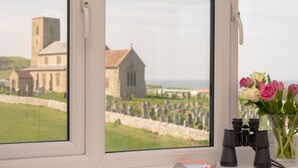 A window with a view of a church and cemetery, a pair of binoculars, and a vase with flowers. - 4 Bed in Cromer (oc-81469) (West Runton)