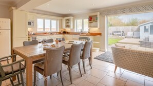 A modern kitchen with white cabinets, a dining area with a wooden table and chairs, and a view of a backyard with outdoor furniture. - 4 Bed in Cromer (oc-81469) (West Runton)