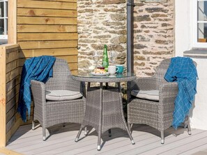 A wicker table with two chairs, a glass of wine, a bottle, and a mug on a deck with a stone wall and wooden panel background. - 1 Bed in Challacombe (oc-81477) (Challacombe)