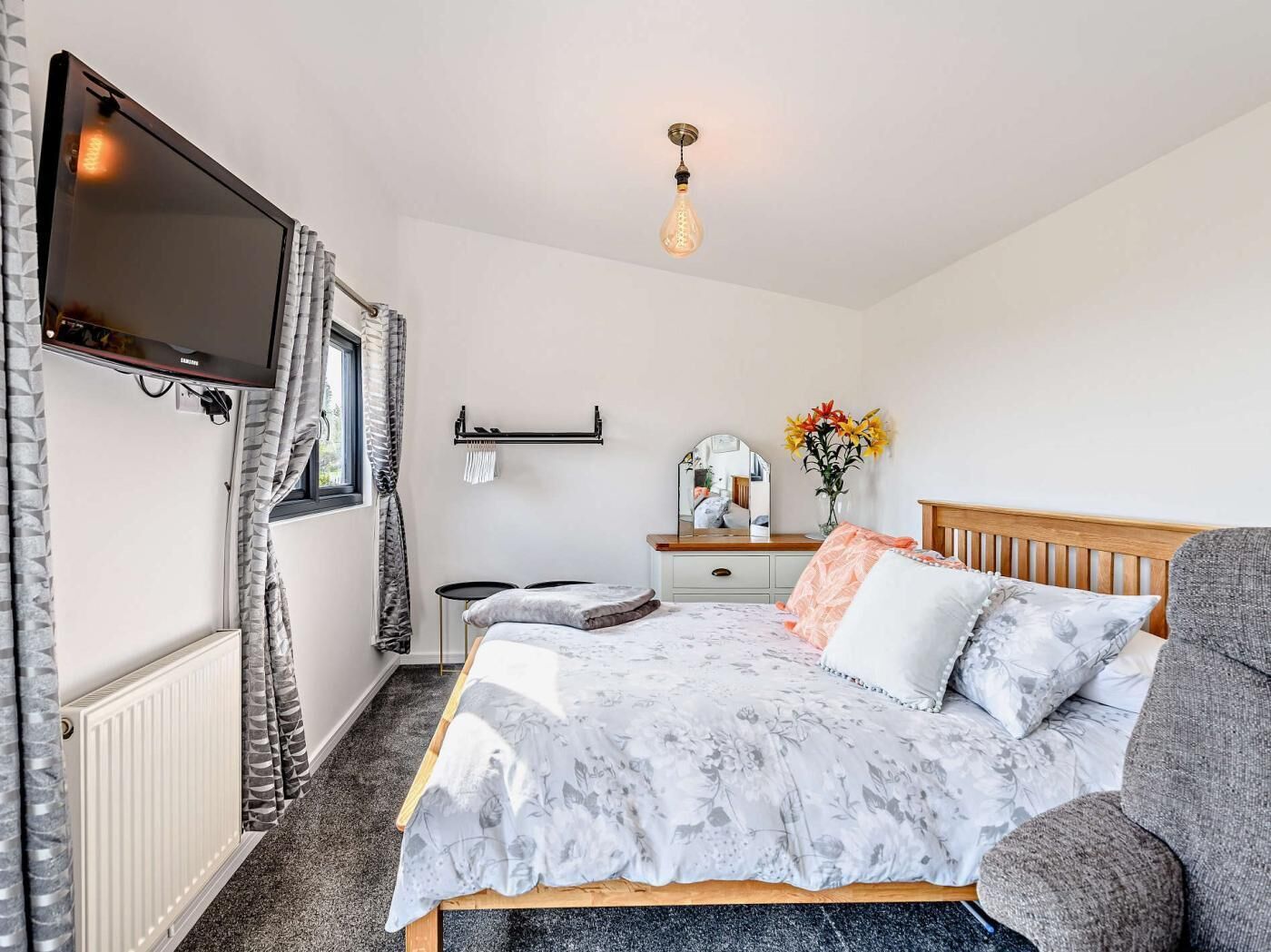 Une chambre à coucher comprenant un lit, une commode, un miroir, un téléviseur fixé au mur et un luminaire suspendu.