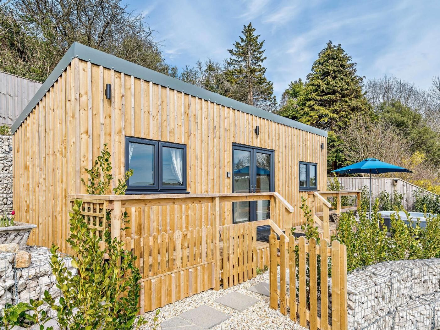 A wooden cabin with a balcony, a stone wall, and a blue umbrella.