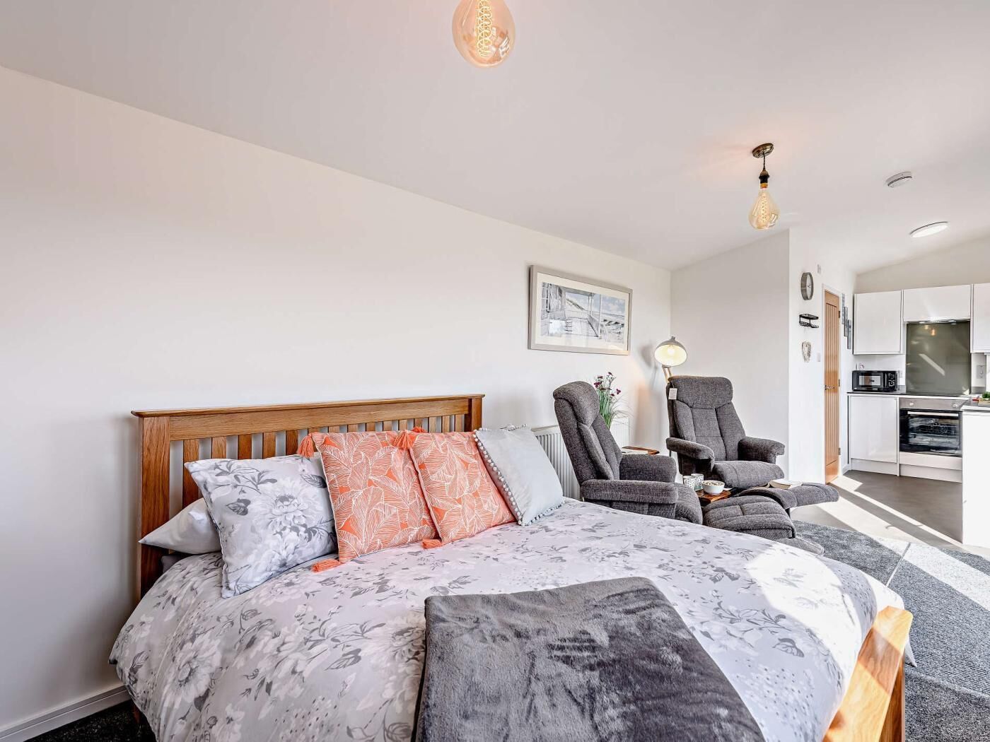 A bedroom with a bed, two armchairs, a desk, and a kitchenette.