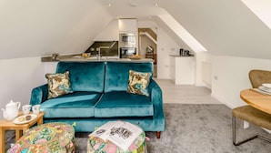 Un accogliente loft con un divano color petrolio, un piccolo tavolino in legno, un poggiapiedi colorato e una cucina sullo sfondo.