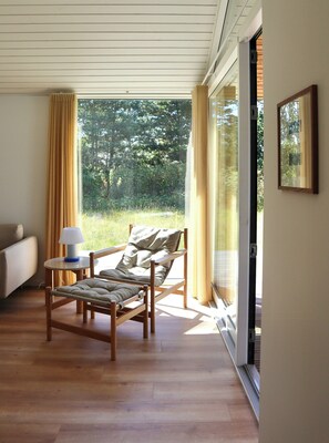 A room with a wooden floor, a single bed, a small table with a lamp, and a view of greenery outside. - New timber house with cozy terrace near the beach (Hals)