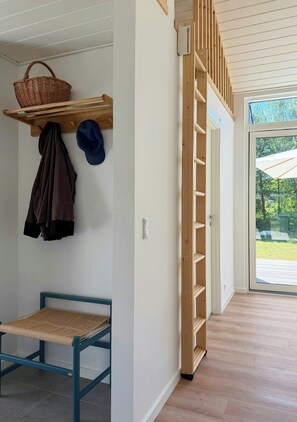 A room with a wooden bench, a wall-mounted shelf with a basket and hats, and a wooden ladder leading to a door. - New timber house with cozy terrace near the beach (Hals)