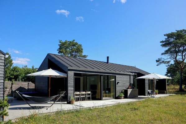 A modern house with a sloped roof, large glass doors, and a deck with outdoor furniture. - New timber house with cozy terrace near the beach (Hals)