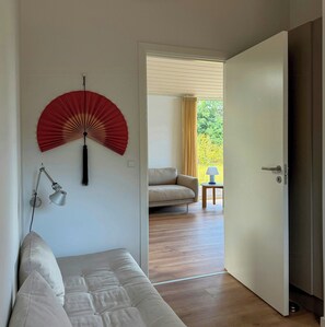 A room with a bed, a red wall fan, a lamp, and a view through an open door to a balcony with a chair and table. - New timber house with cozy terrace near the beach (Hals)