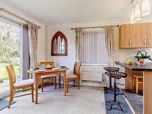 A dining area with a table and chairs, a kitchen with wooden cabinets, and a window with curtains. - 1 Bed in North Walsham (oc-82143) (North Walsham)