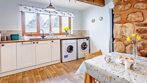 A kitchen with wooden cabinets, a washing machine, a table with a tablecloth, and a stone wall. - 1 Bed in Watchet (oc-81240) (Washford)