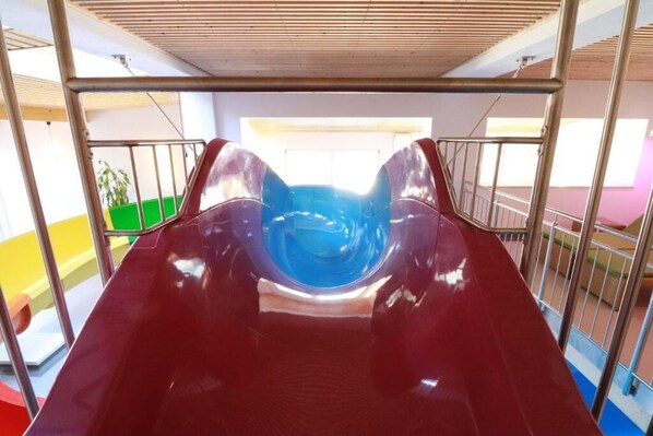 Un tobogán rojo con una sección central azul, rodeado de barandales metálicos, en un área de juegos interior bien iluminada.