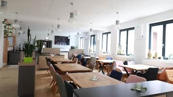 A modern dining area with multiple tables and chairs, large windows, and pendant lighting.