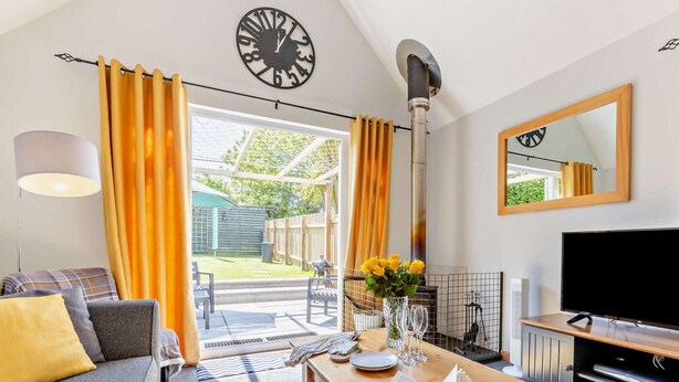 A modern living room with a sofa, a dining table with a book and glasses, a TV, and a large window with curtains. - 2 Bed in Bude (oc-82163) (Bude)