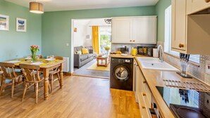 A compact kitchen with a washing machine, a dining area with wooden chairs, and a view to a living room. - 2 Bed in Bude (oc-82163) (Bude)