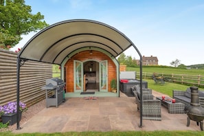 A covered outdoor seating area with a barbecue, a hot tub, and a view of a house in the background.