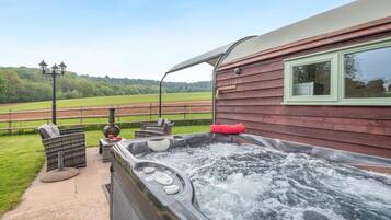 Un bain à remous avec vue sur un champ verdoyant, un bâtiment en bois et du mobilier d’extérieur.