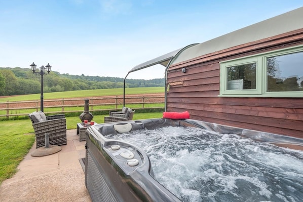 A hot tub with a view of a green field, a wooden building, and outdoor furniture.