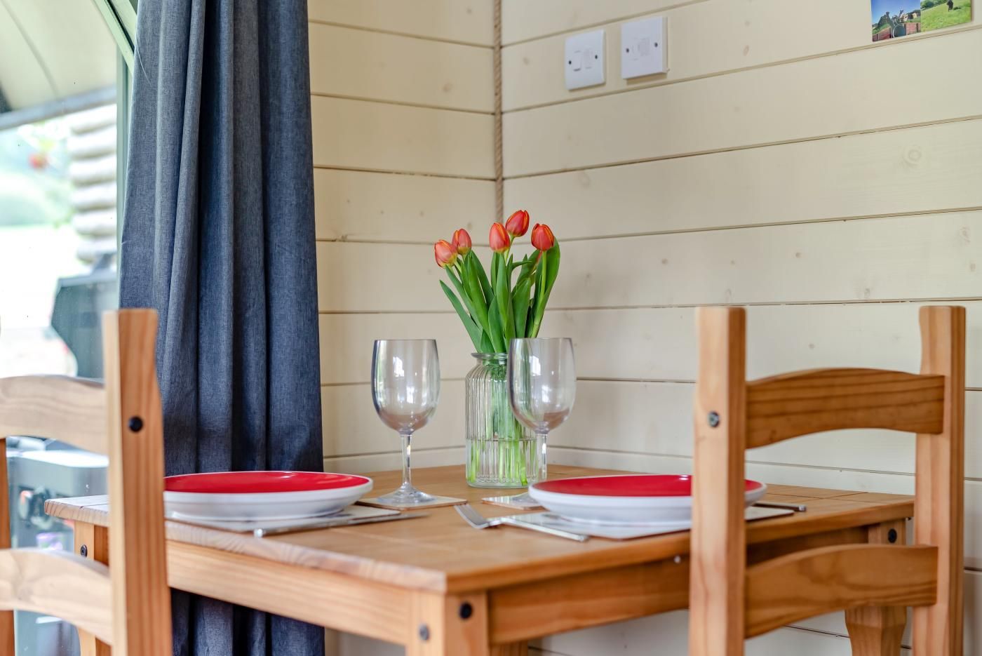 Een houten tafel met twee wijnglazen en een vaas tulpen, voor een wand met panelen en een gordijn.
