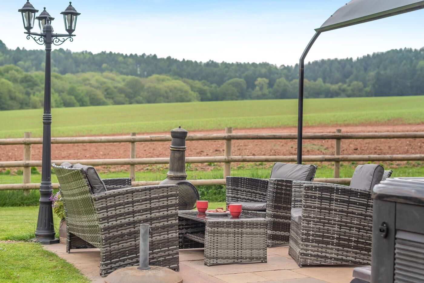 Mobilier d’extérieur en osier, comprenant une table et des chaises, un lampadaire et une vue sur un champ verdoyant.