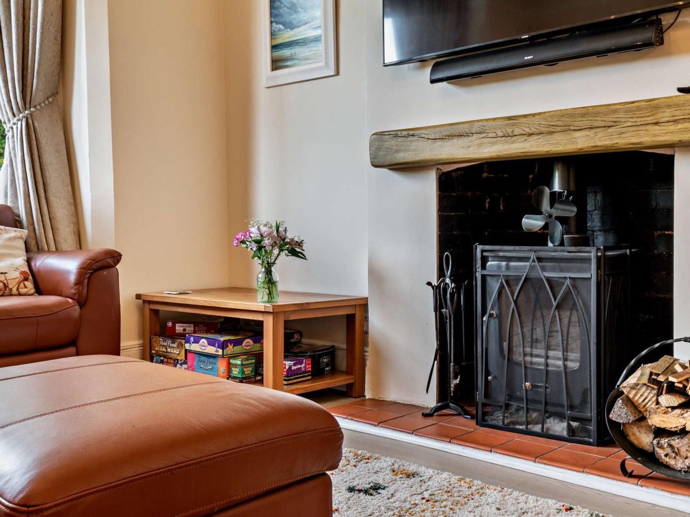 A living room with a leather sofa, a small wooden table with a vase of flowers, a fireplace with a log basket, and a television mounted above the fireplace.