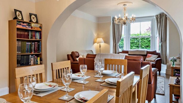 Un coin repas avec une table en bois dressée pour quatre personnes, une bibliothèque, un canapé en cuir et un lustre.