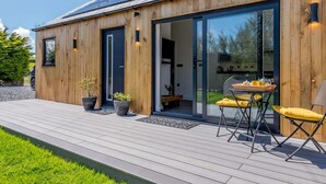 A modern wooden building with a solar panel roof, a glass door entrance, and a deck with outdoor furniture. - 1 Bed in Woolacombe (oc-82341) (Woolacombe)