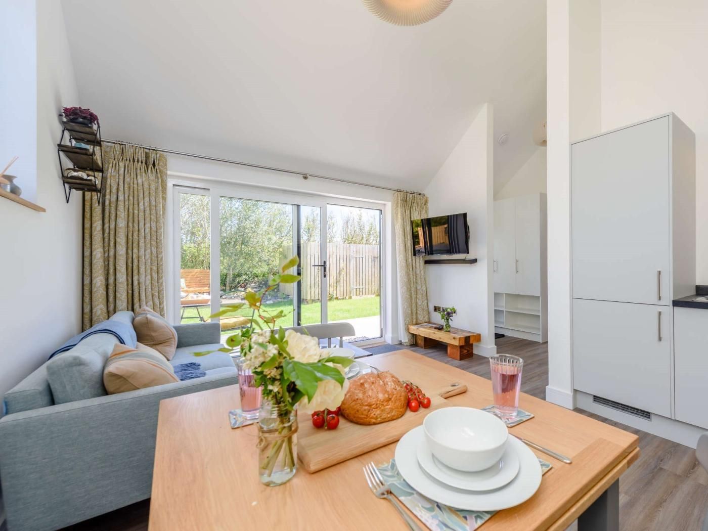 A modern living room with a sofa, a dining table set with plates, a vase with flowers, and a kitchen area with cabinets and a TV.