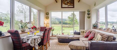 A spacious room with a dining area, a sofa set, and large windows offering a view of the countryside.