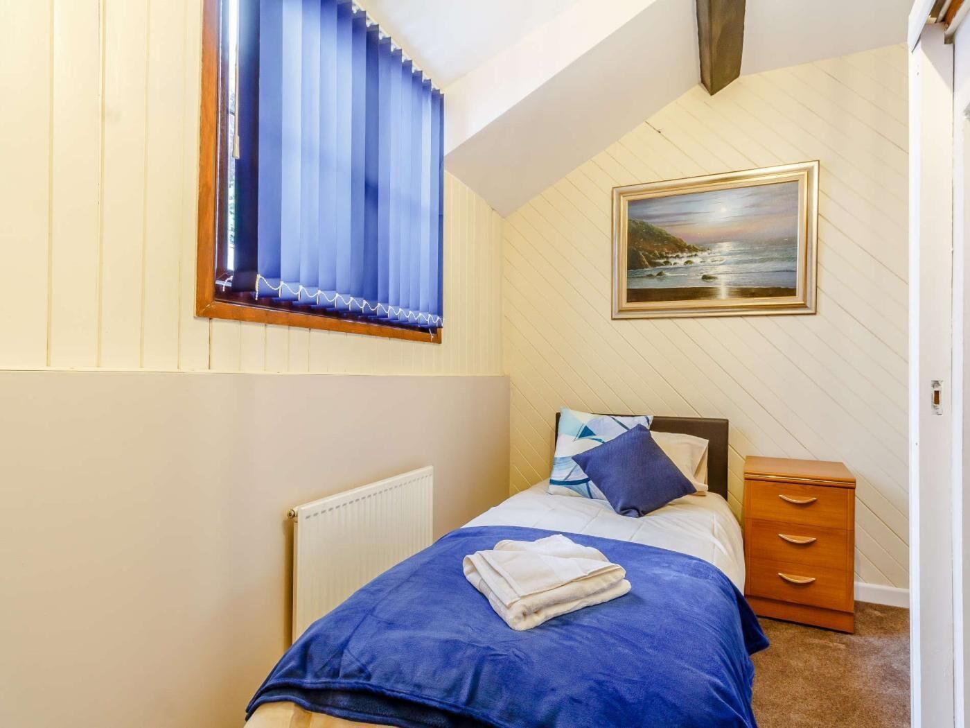 A bedroom with a bed, a nightstand, a radiator, a window with blinds, and a painting on the wall.