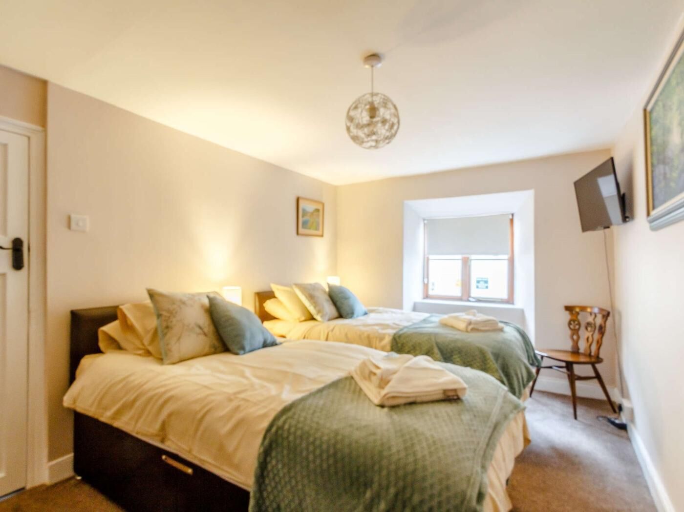 A bedroom with two beds, a window, a wall-mounted TV, and a chandelier.