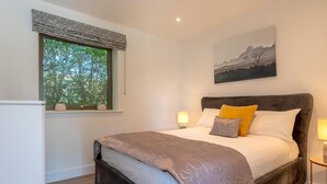 Een slaapkamer met een groot bed, een raam met uitzicht op bomen, een schilderij aan de muur en een nachtkastje met een lamp.