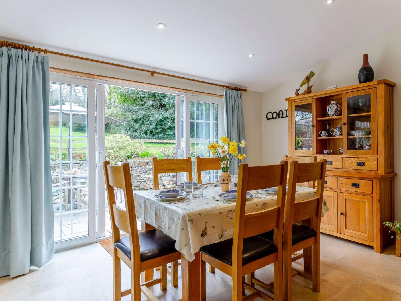 A dining area with a table set for four, wooden chairs, a cabinet with glass doors, and a view of a garden through large windows.