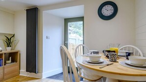 Un coin repas avec une table ronde, des chaises blanches, une horloge au mur et une vue sur l’extérieur, visible à travers une porte ouverte.
