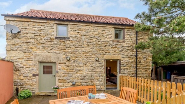 Un bâtiment en pierre avec un toit en tuiles, une table en bois avec des chaises et une clôture en bois.